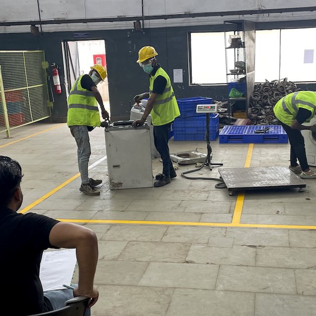 Two workers wearing yellow hard hats and safety vests dismantling a white appliance in a workshop while a supervisor observes