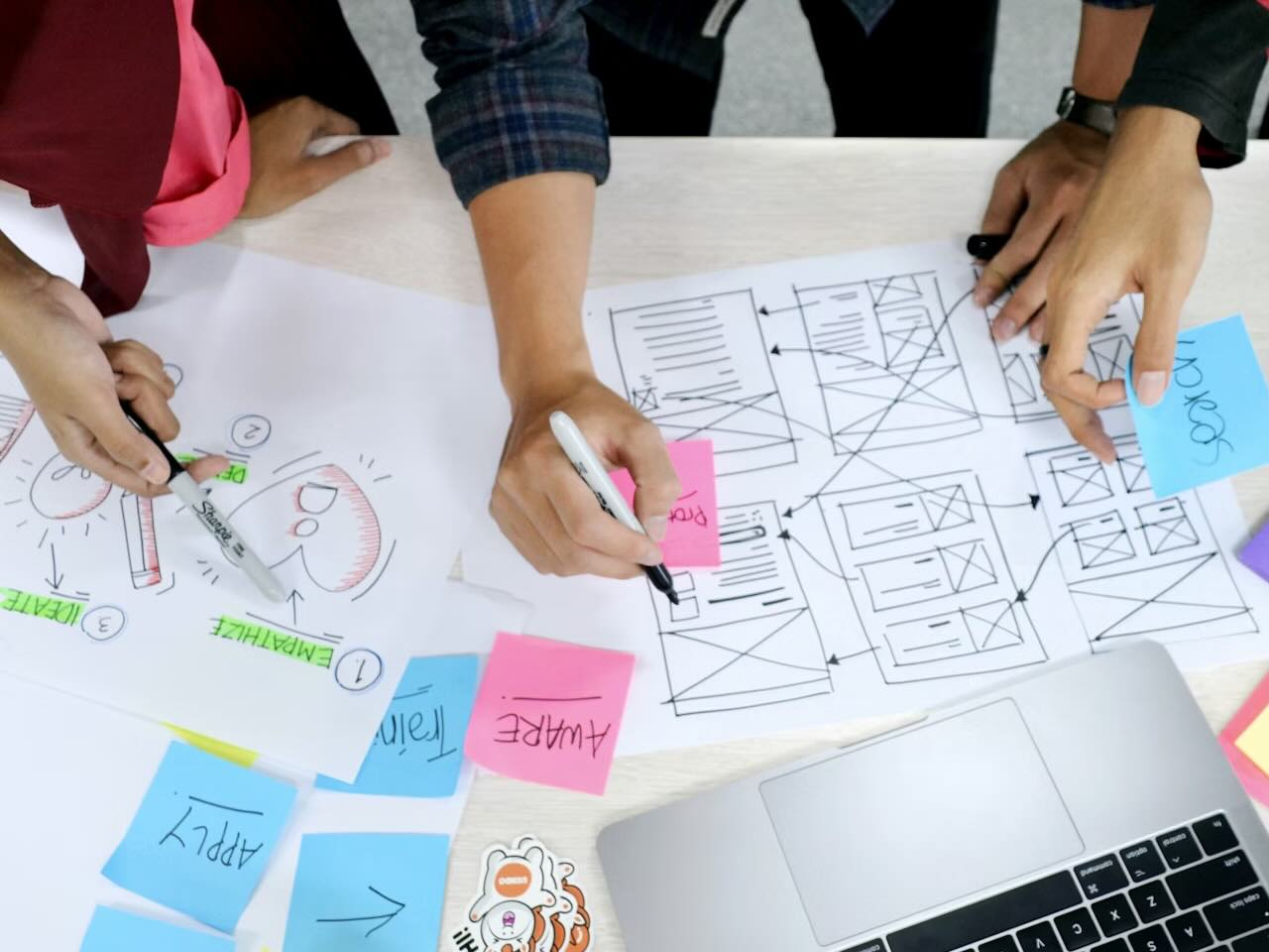 A desk with a MacBook, sticky notes, and paper, with multiple pairs of hands sketching UX wireframes and illustrations on the paper sheets