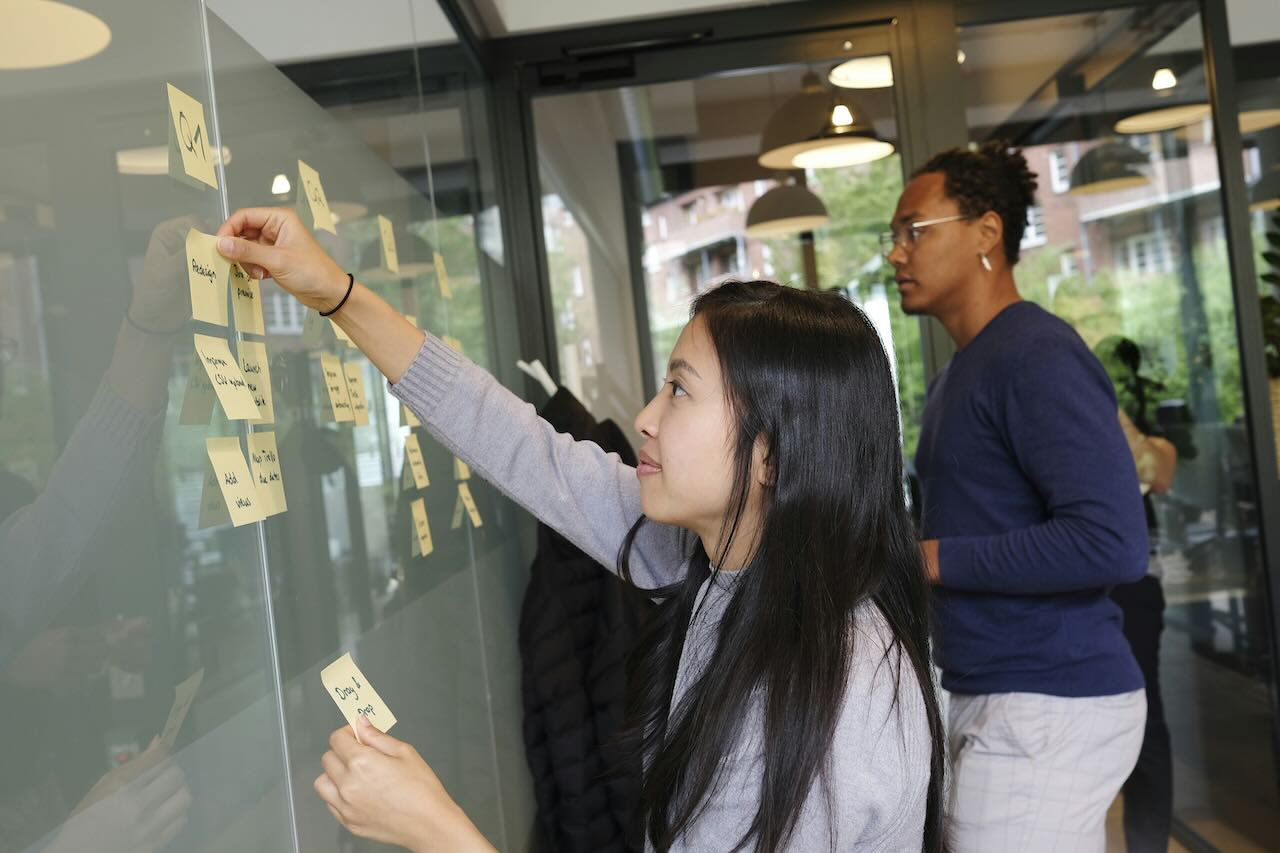 Two people place sticky notes on a whiteboard made out of glass